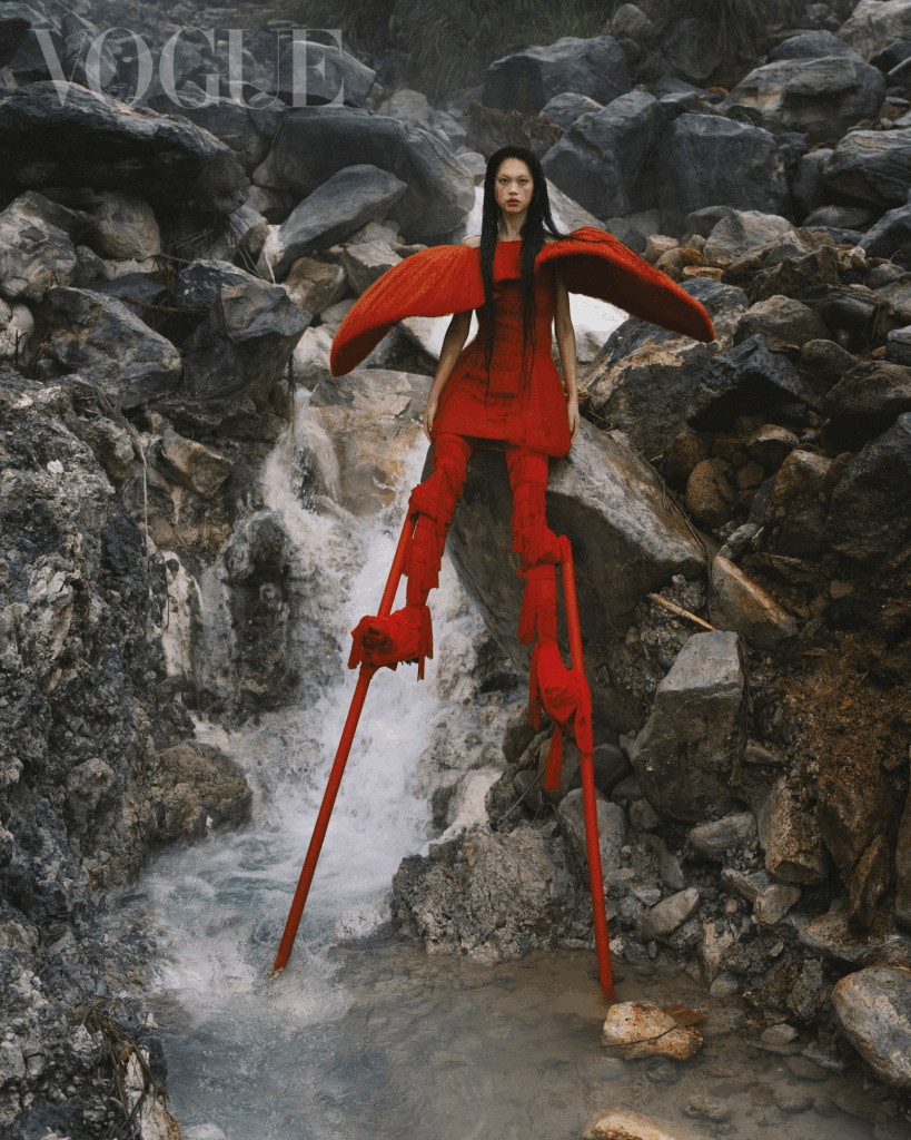 Small model in bright red winged cape and dress with stillt like shoes, posing over dangerous looking small fast running stony stream